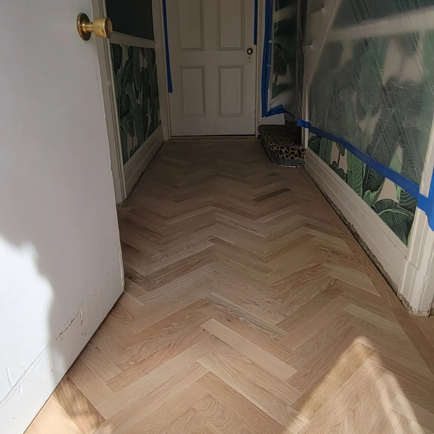 A hallway featuring light wood flooring laid in a herringbone pattern, with patterned wallpaper and blue painter's tape.