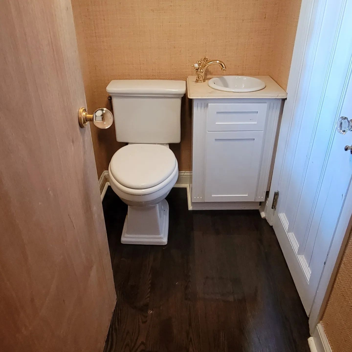 A small powder room with a white toilet, white vanity with a gold faucet, tan walls, and dark wood-look flooring.