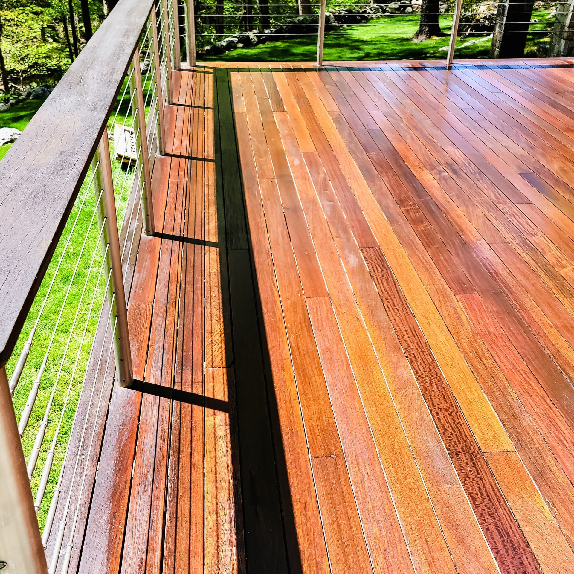 A wooden deck with reddish-brown planks and a stainless steel cable railing overlooking a green, sunlit lawn.