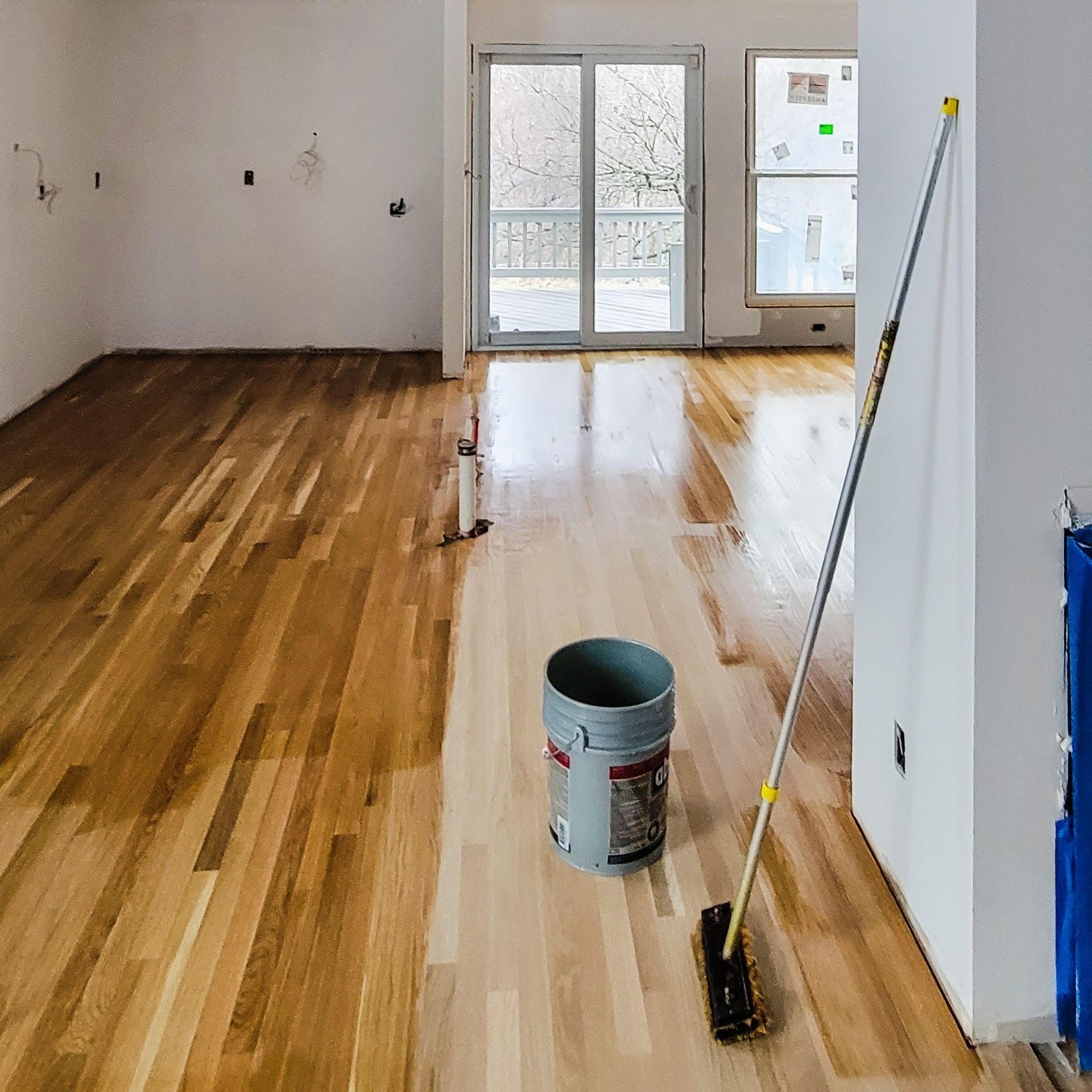 An interior view of a room with partially finished hardwood floors, a bucket, and a long-handled applicator tool.