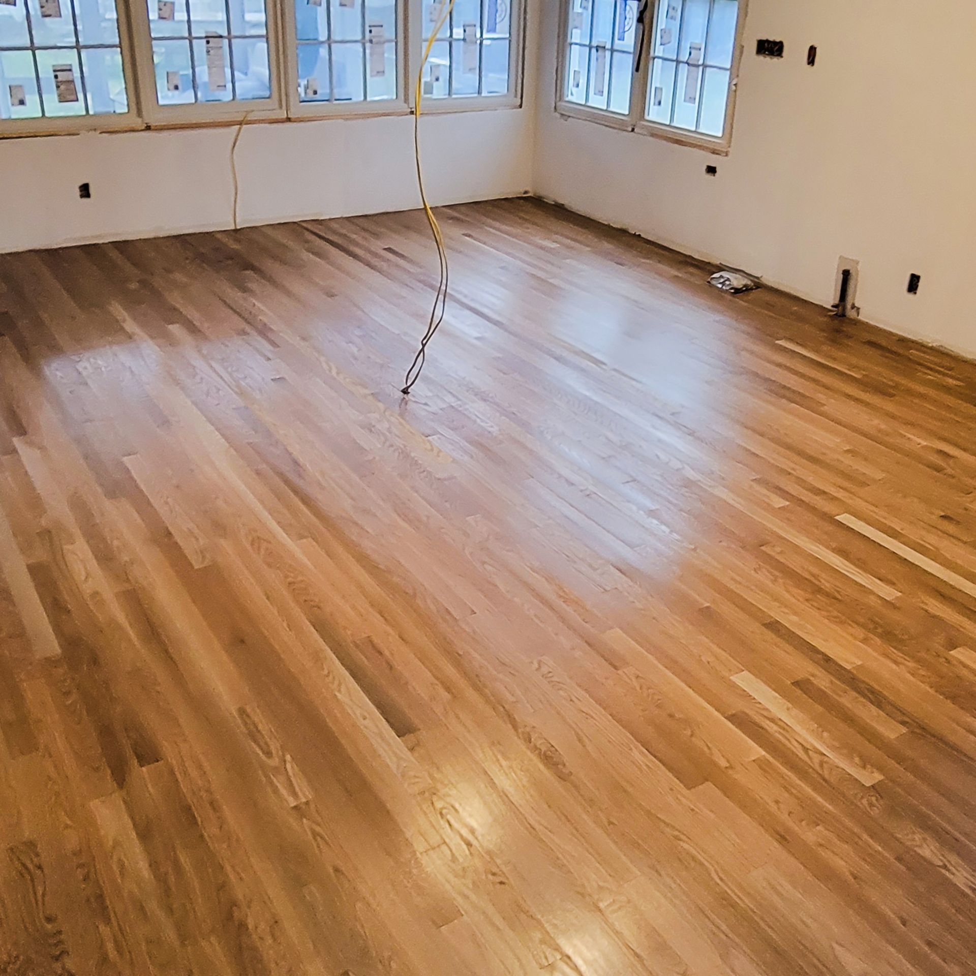 Newly installed light-toned wood flooring in a room with white walls, multiple windows, and a hanging electrical wire.