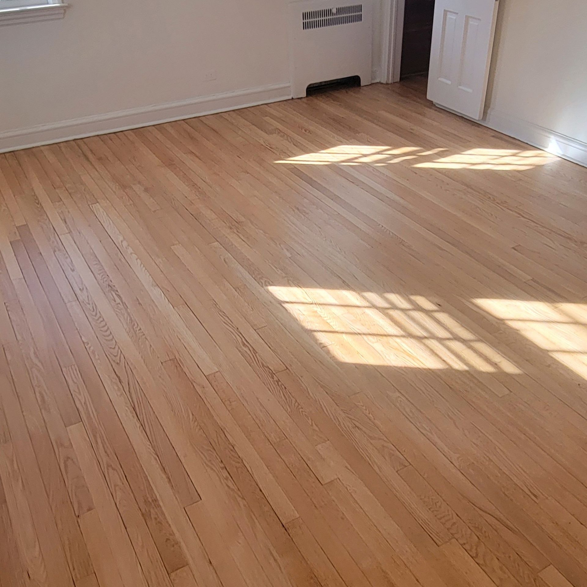 A room with light-colored, recently refinished hardwood floors, a white wall, a floor vent, and a white interior door.