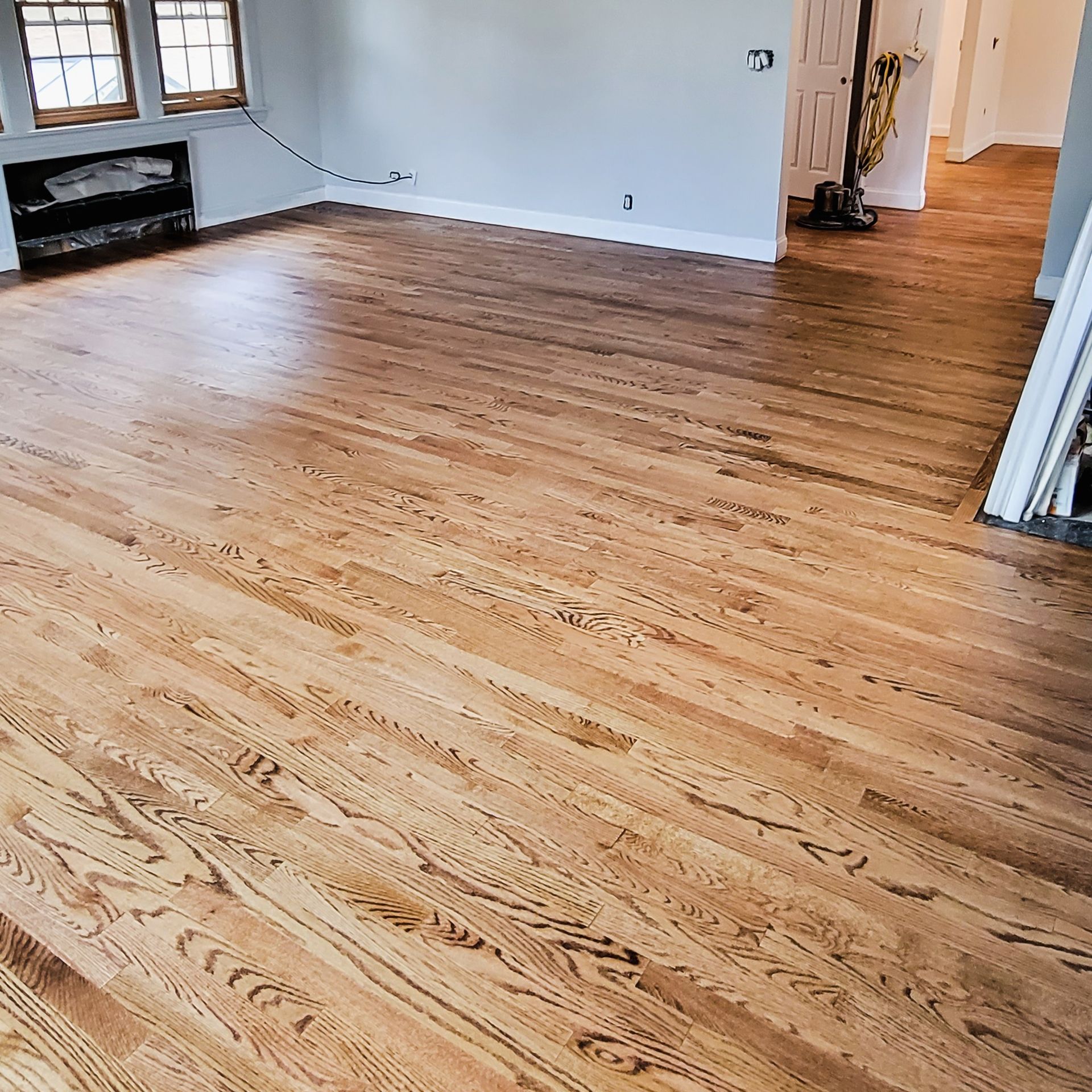 A bright room with newly refinished, light-colored hardwood floors, white baseboards, and windows on a gray wall.