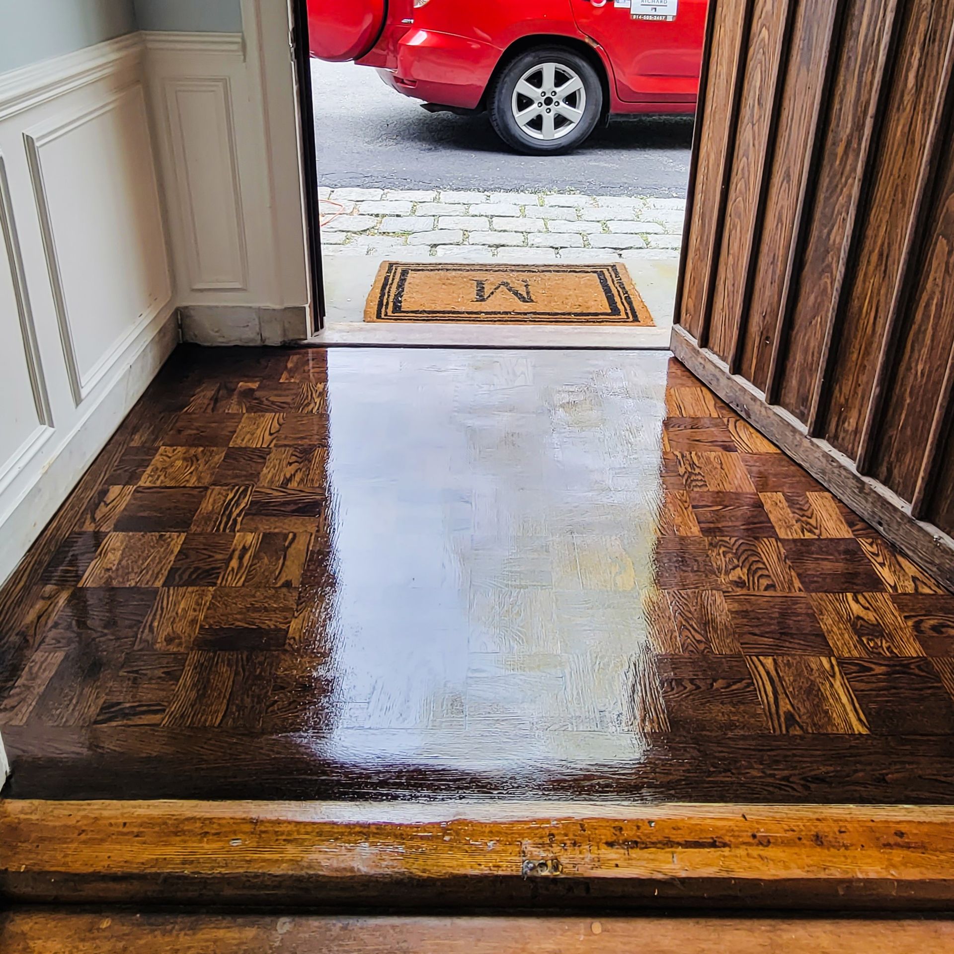 Dark wood parquet flooring leads to an open doorway with a welcome mat, revealing a red car parked outside.