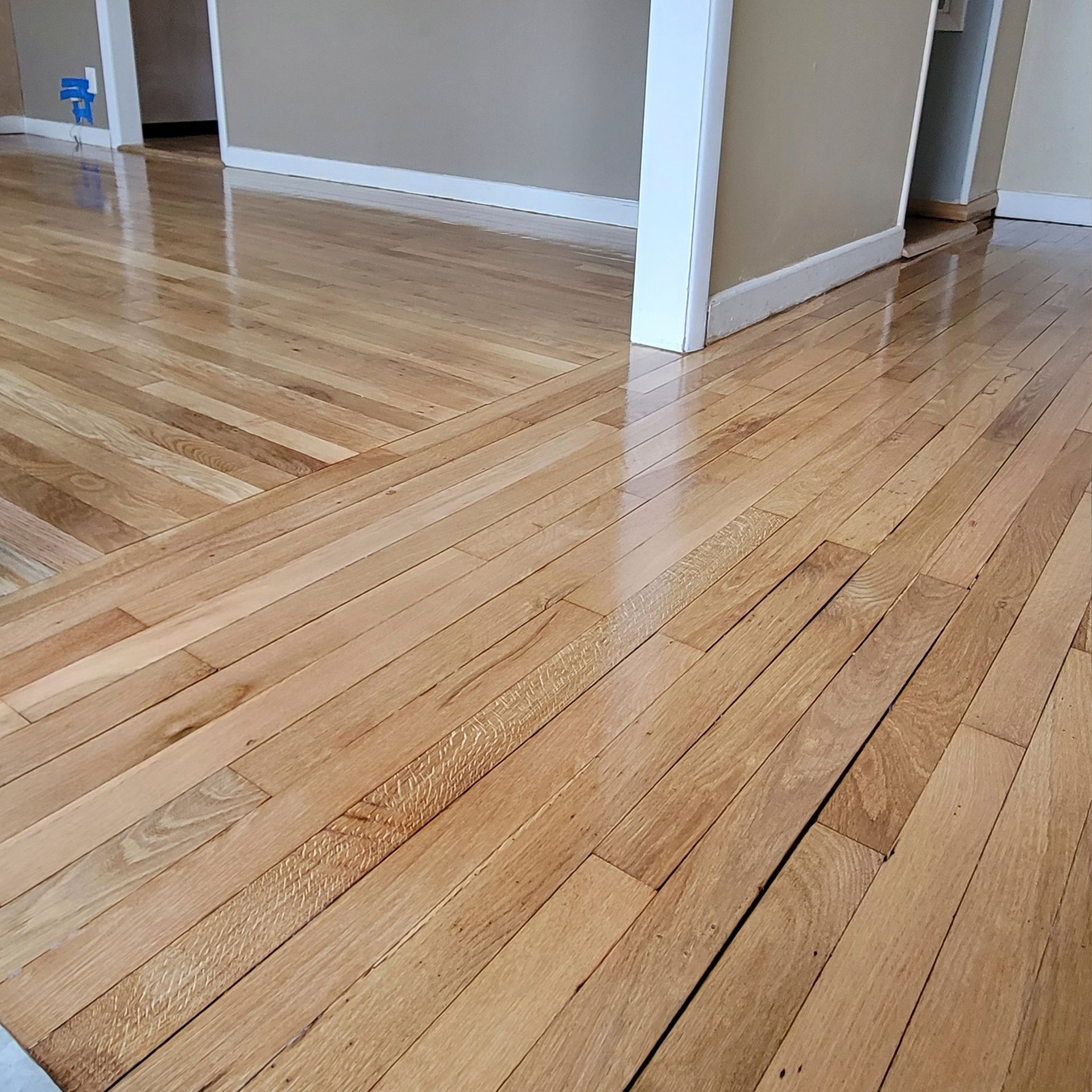 Light-colored hardwood flooring meeting at a threshold with white baseboards and neutral walls in an indoor space.