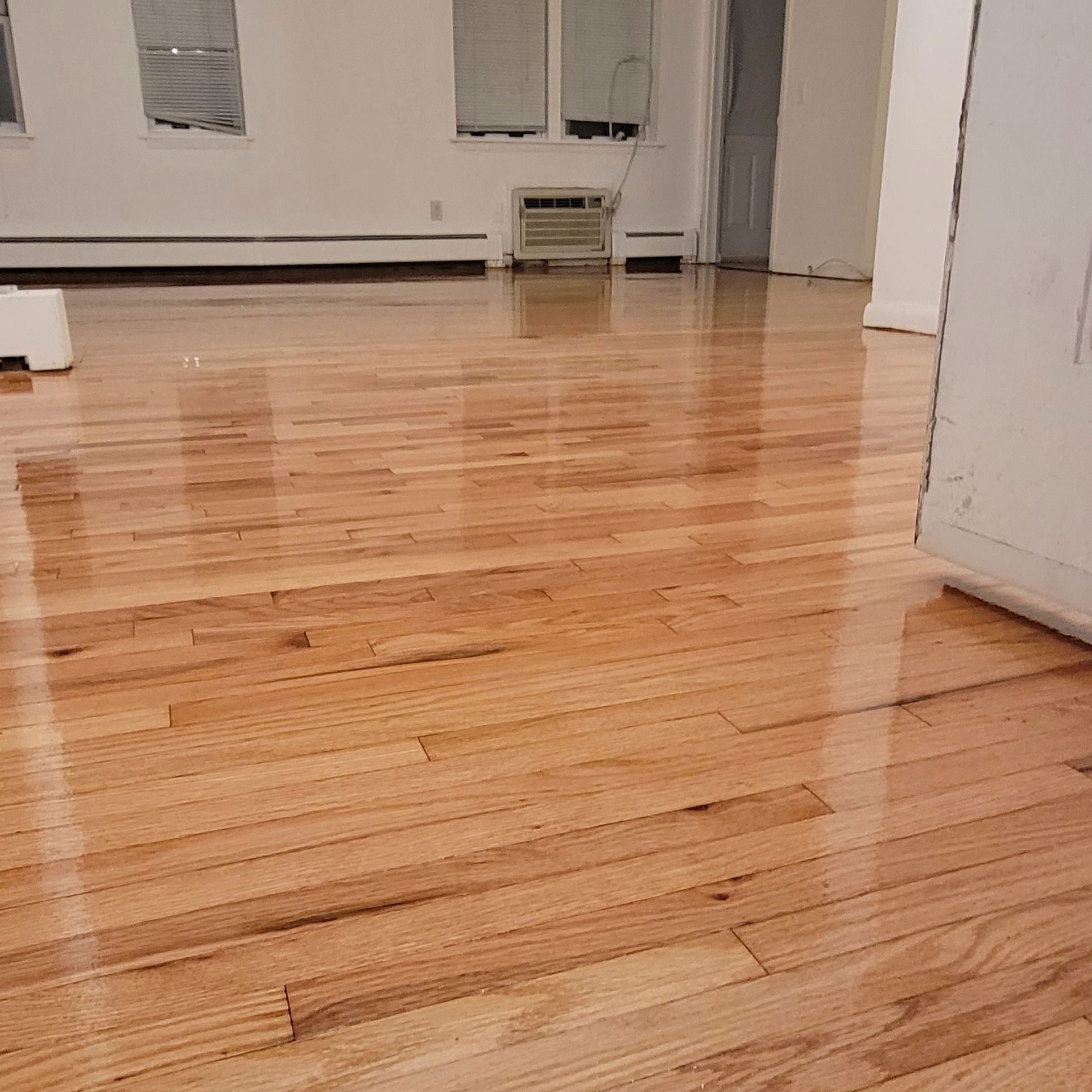 A light-colored, glossy hardwood floor in an empty room with white walls and a window air conditioning unit.