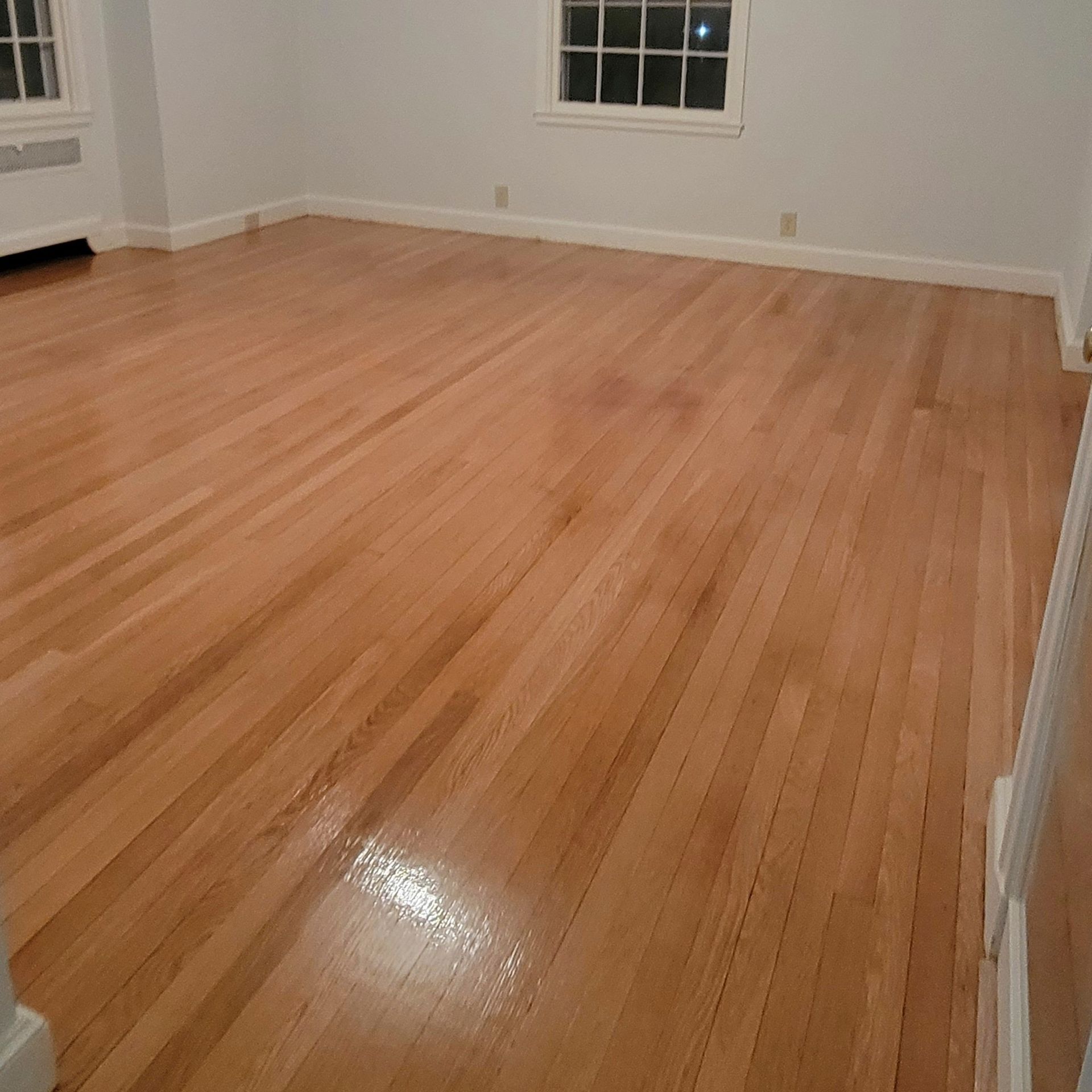 A bright, empty room with polished light-wood floors and white walls featuring a small window and floor vents.