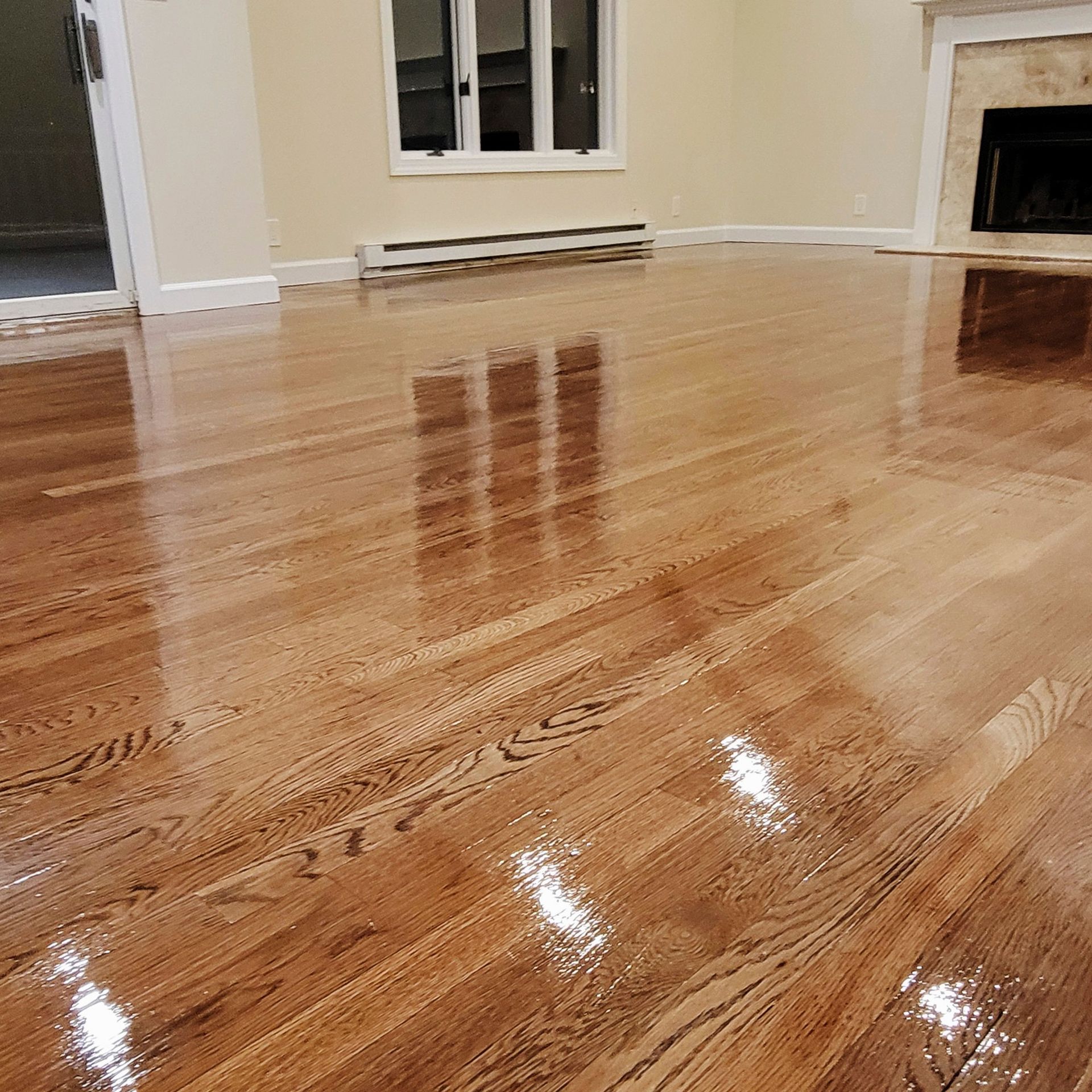 A polished hardwood floor in a room featuring light walls, a window, a white baseboard heater, and a fireplace.