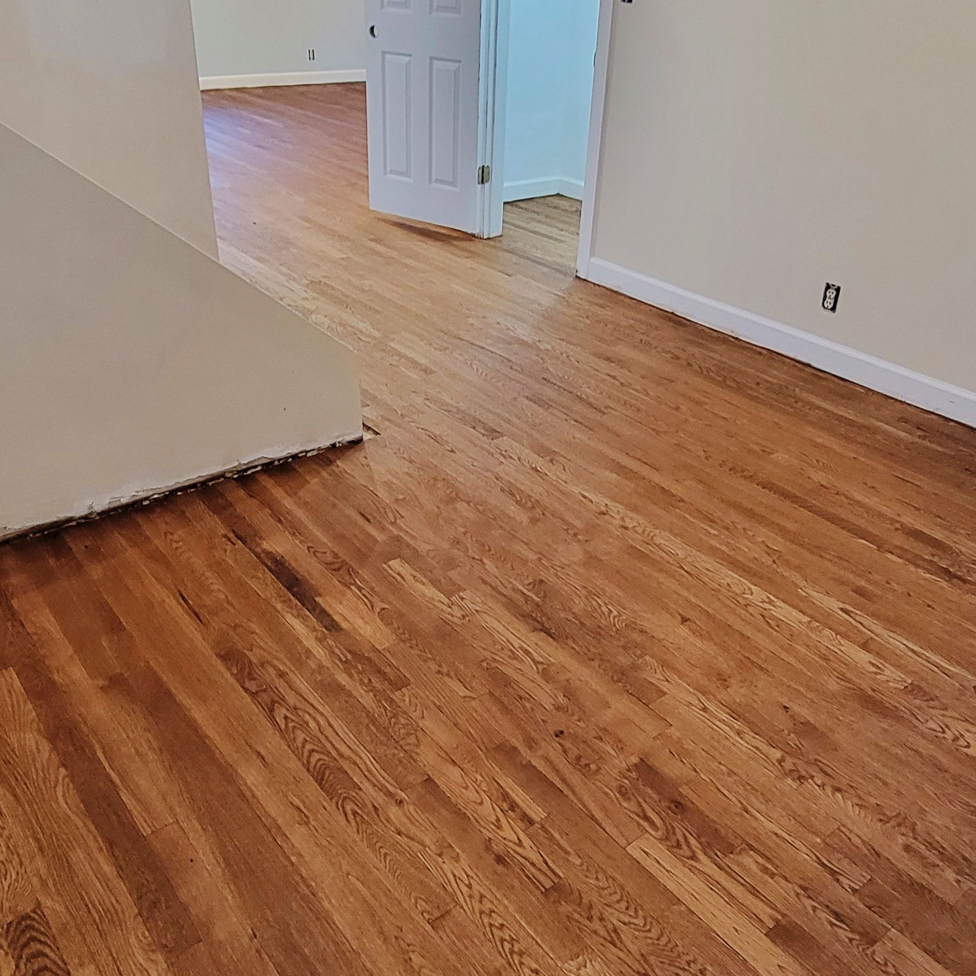 A room with polished, light-brown hardwood flooring, beige walls, white trim, and an open white door leading to a hallway.