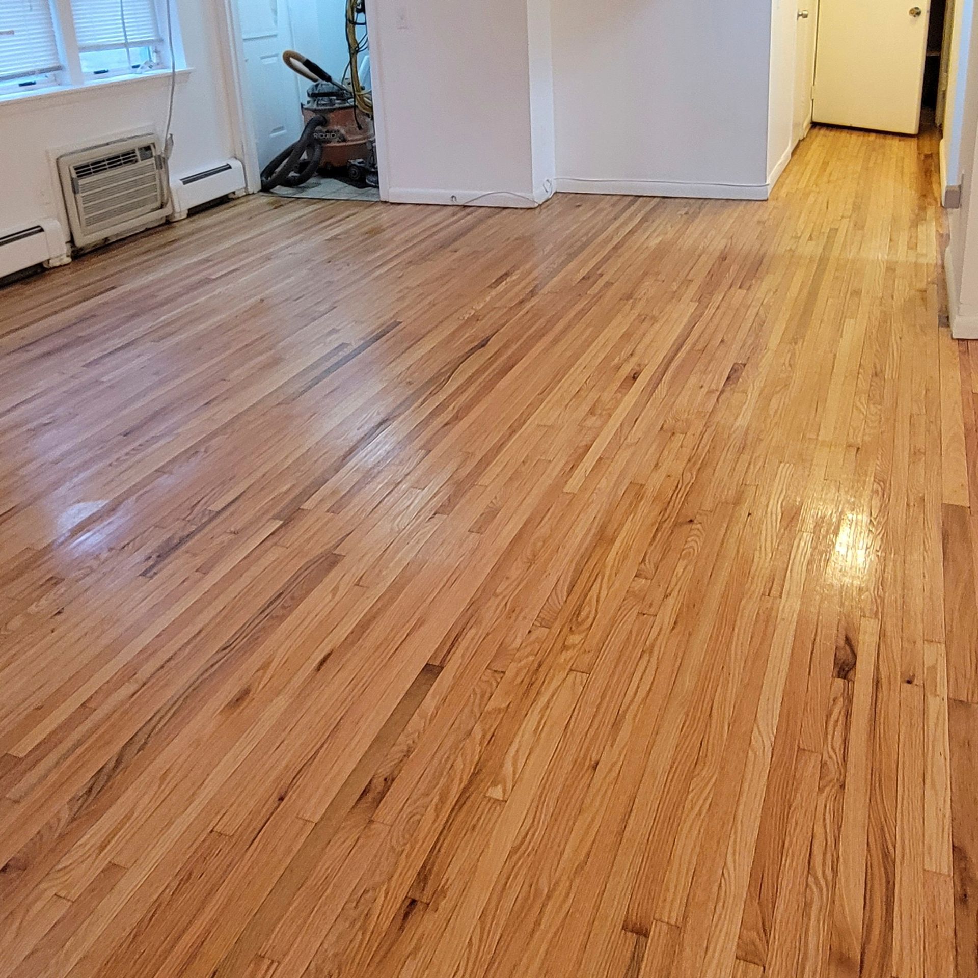 A view of a newly refinished, light-colored hardwood floor in an empty room with white walls and a white radiator.