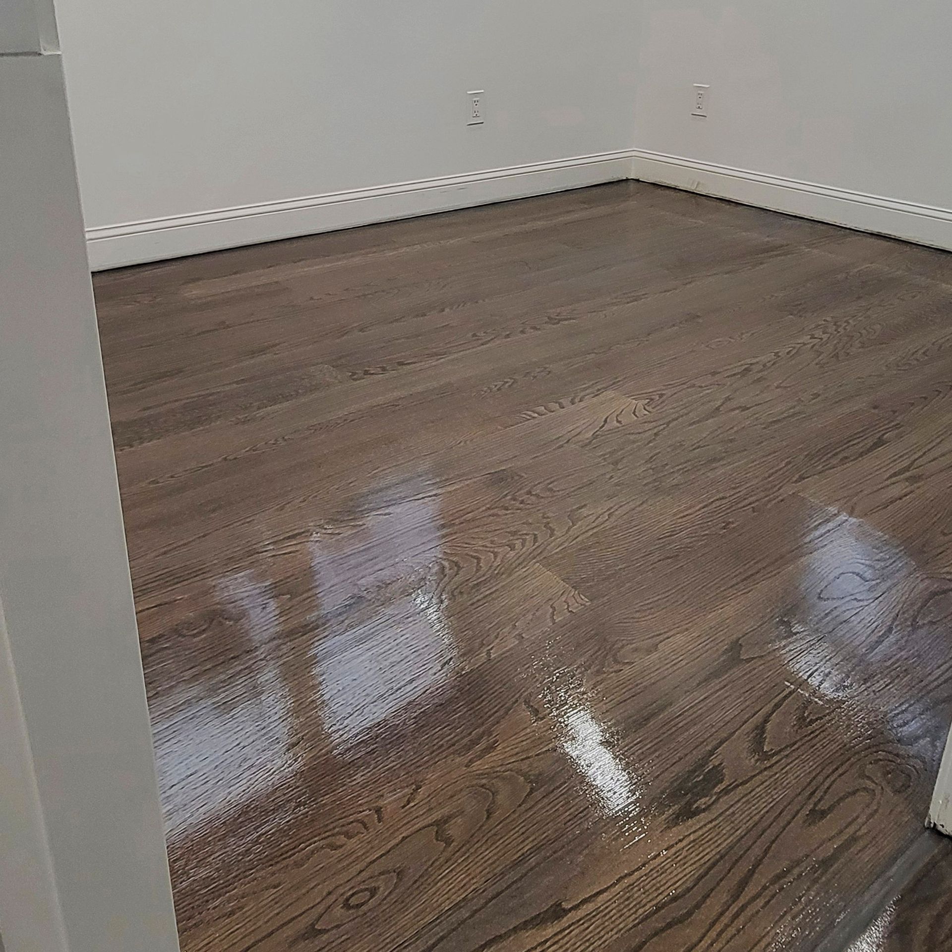A room with glossy, medium-brown stained hardwood floors and off-white baseboards against plain walls.