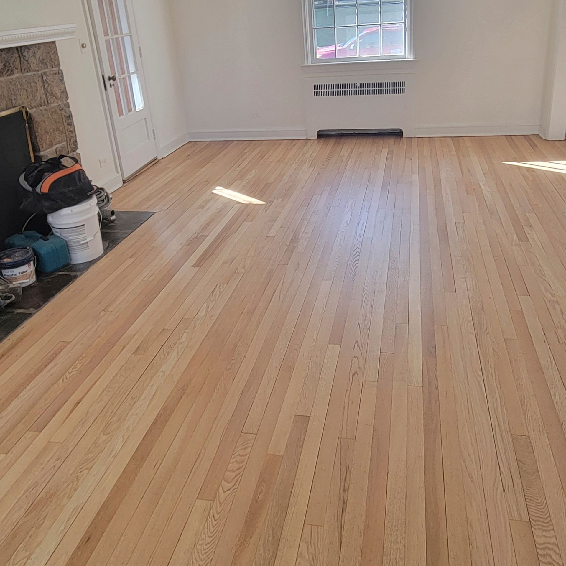 A freshly sanded, light-toned hardwood floor in a room with a white wall, window, and a fireplace corner.