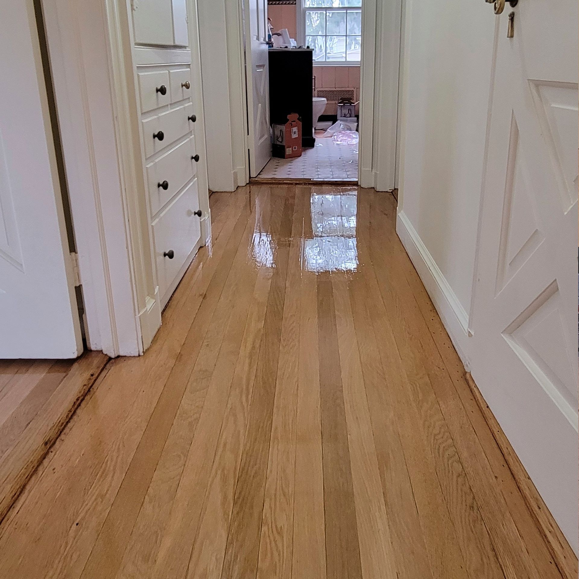 A bright hallway with light wood flooring showing a clear reflection of the ceiling, leading toward a bathroom.
