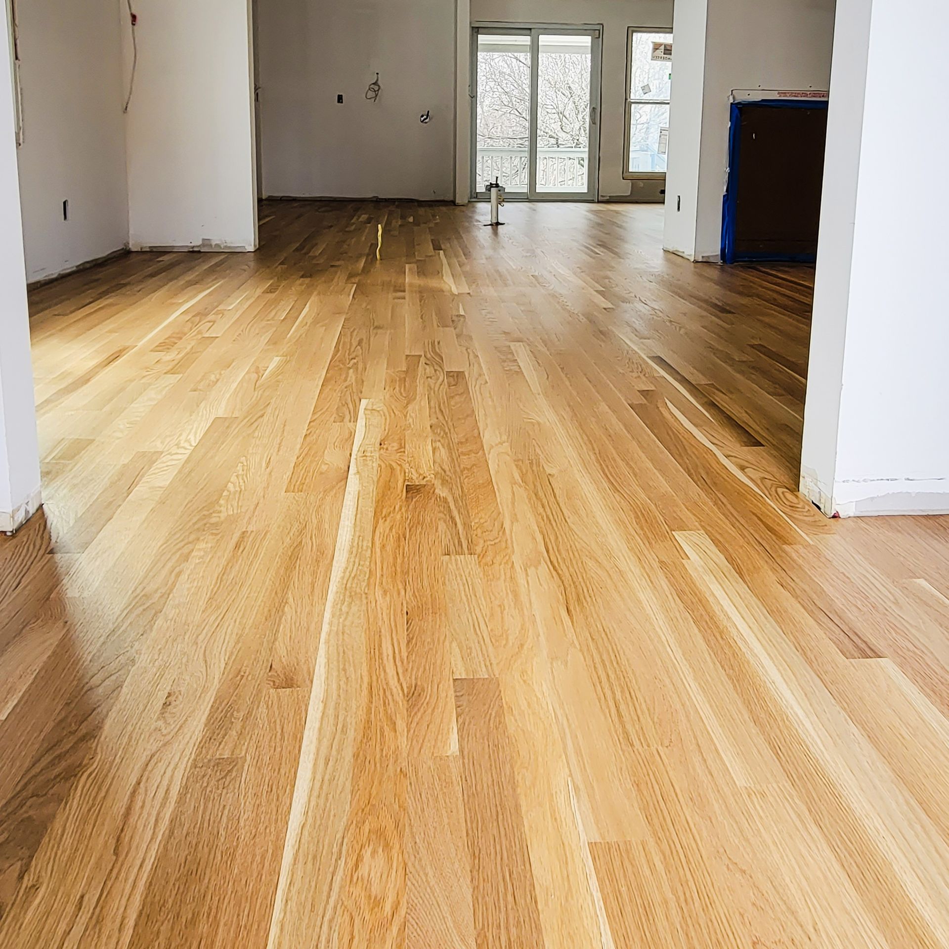 Newly installed light-colored hardwood flooring stretches through an open-concept room with white walls.