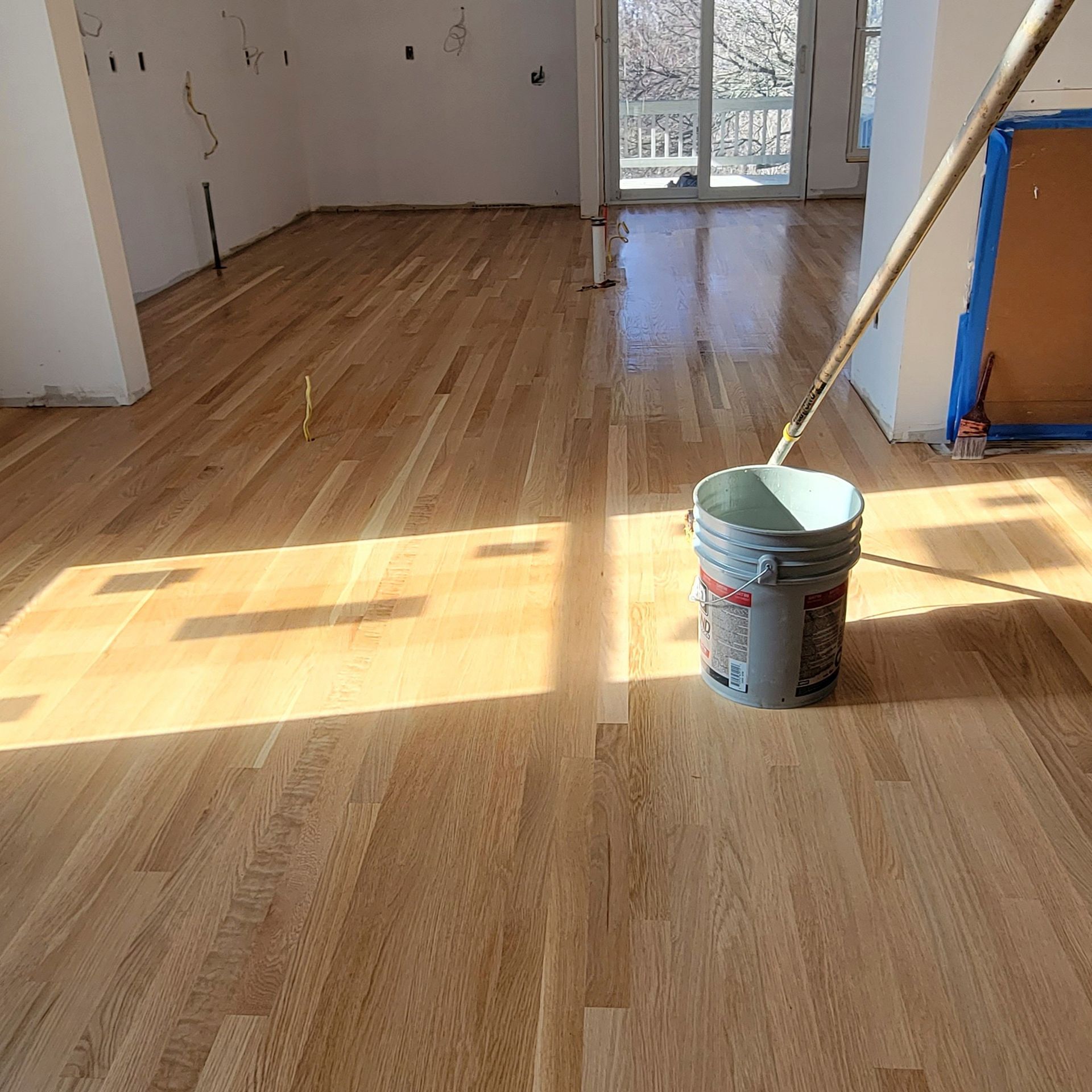 Freshly finished light-colored hardwood floors in an interior room with a bucket and applicator tool.