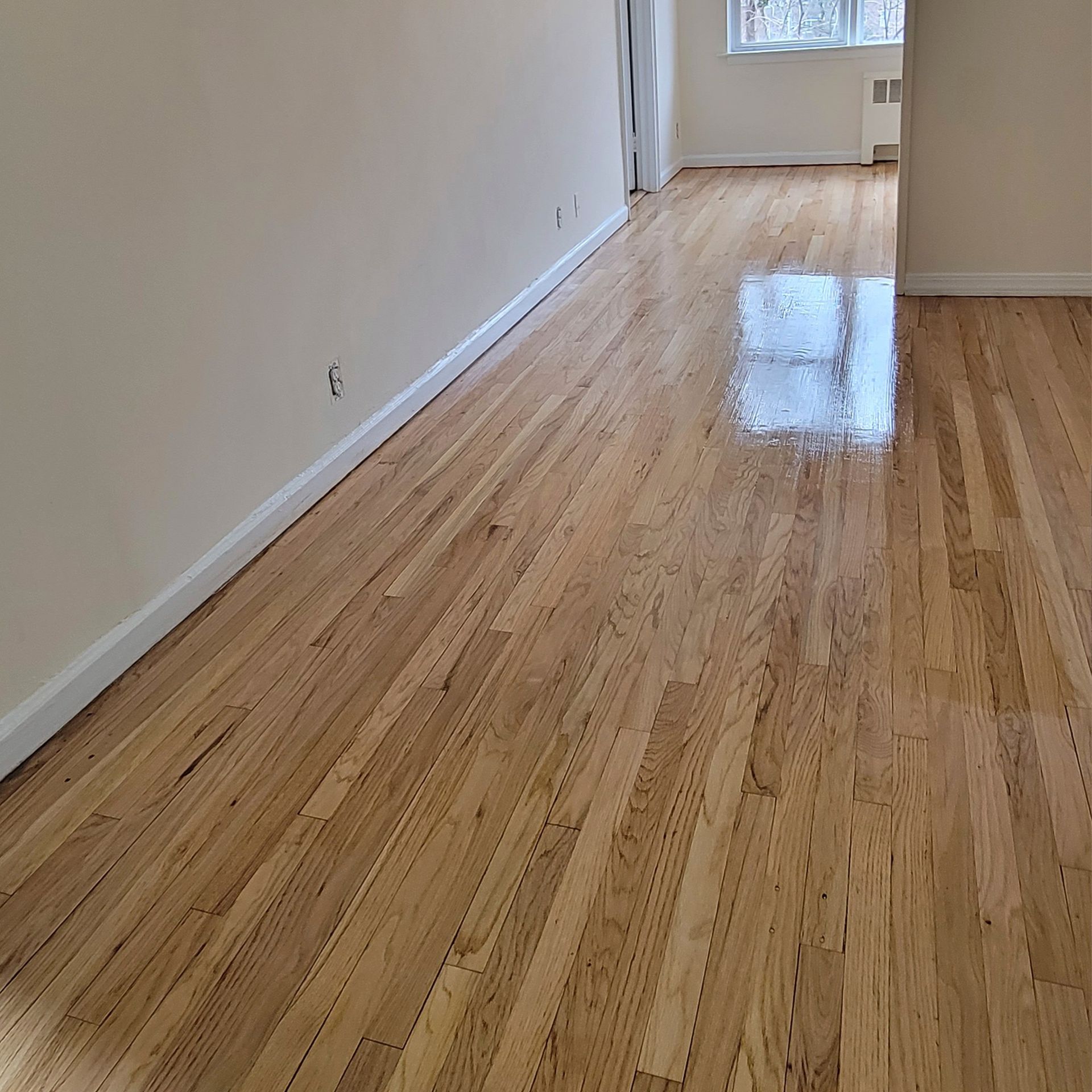 A hallway with light-colored hardwood floors and beige walls, leading toward a window at the far end.