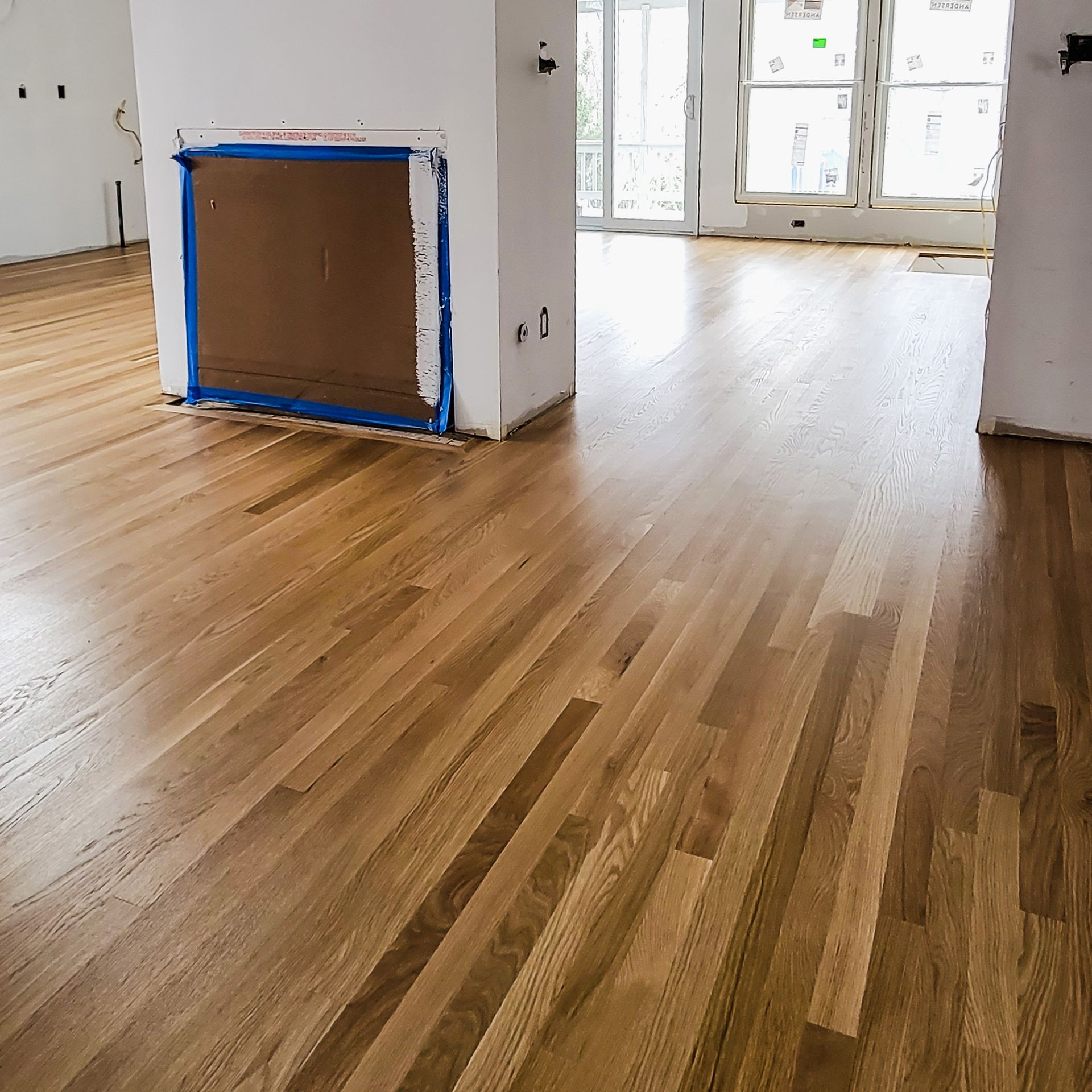Newly installed light brown hardwood flooring in an unfinished room with a covered fireplace and large windows.