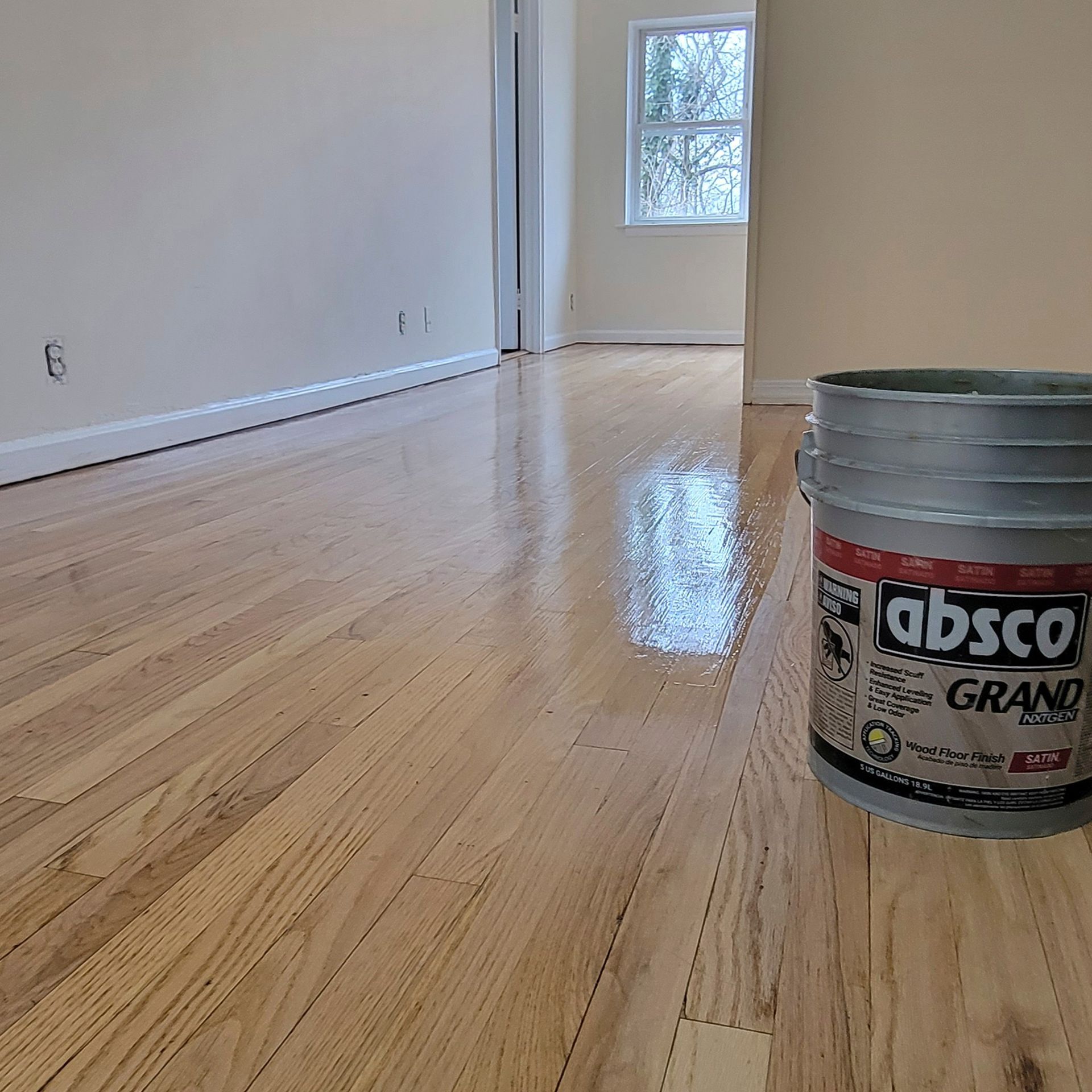 A bucket of Absco Grand floor finish sits on a newly polished, light-colored hardwood floor in a room.
