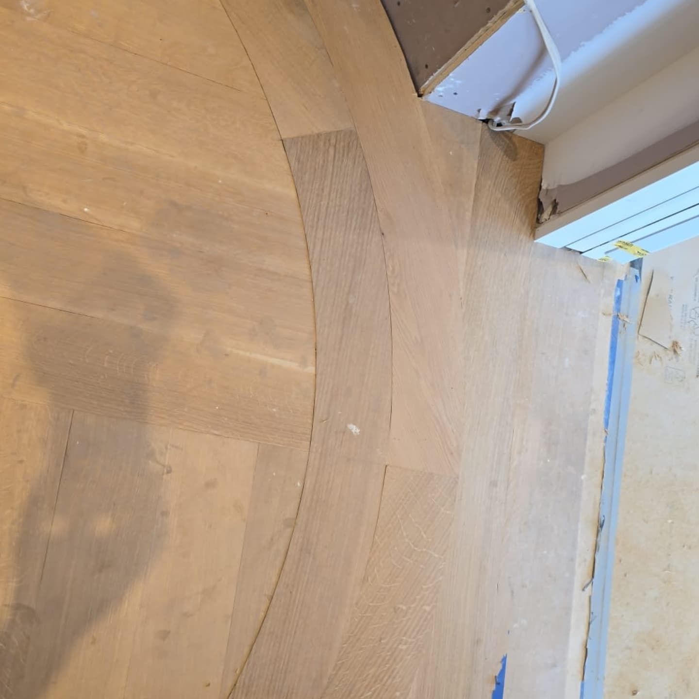 Light-colored wooden floor planks installed in a curved pattern near a doorway with an exposed wall and cable.