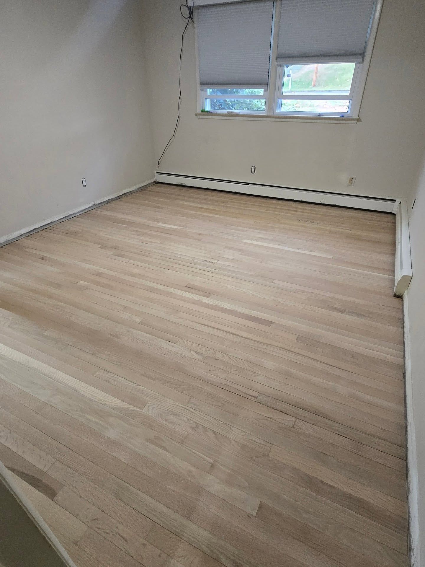 An empty room featuring light-colored hardwood flooring, cream-colored walls, a window with blinds, and baseboard heating.