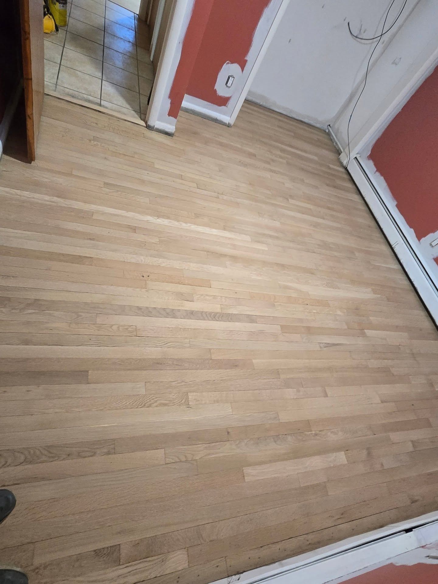 Newly sanded, light-colored hardwood floor in a room with partially painted reddish walls and white baseboards.
