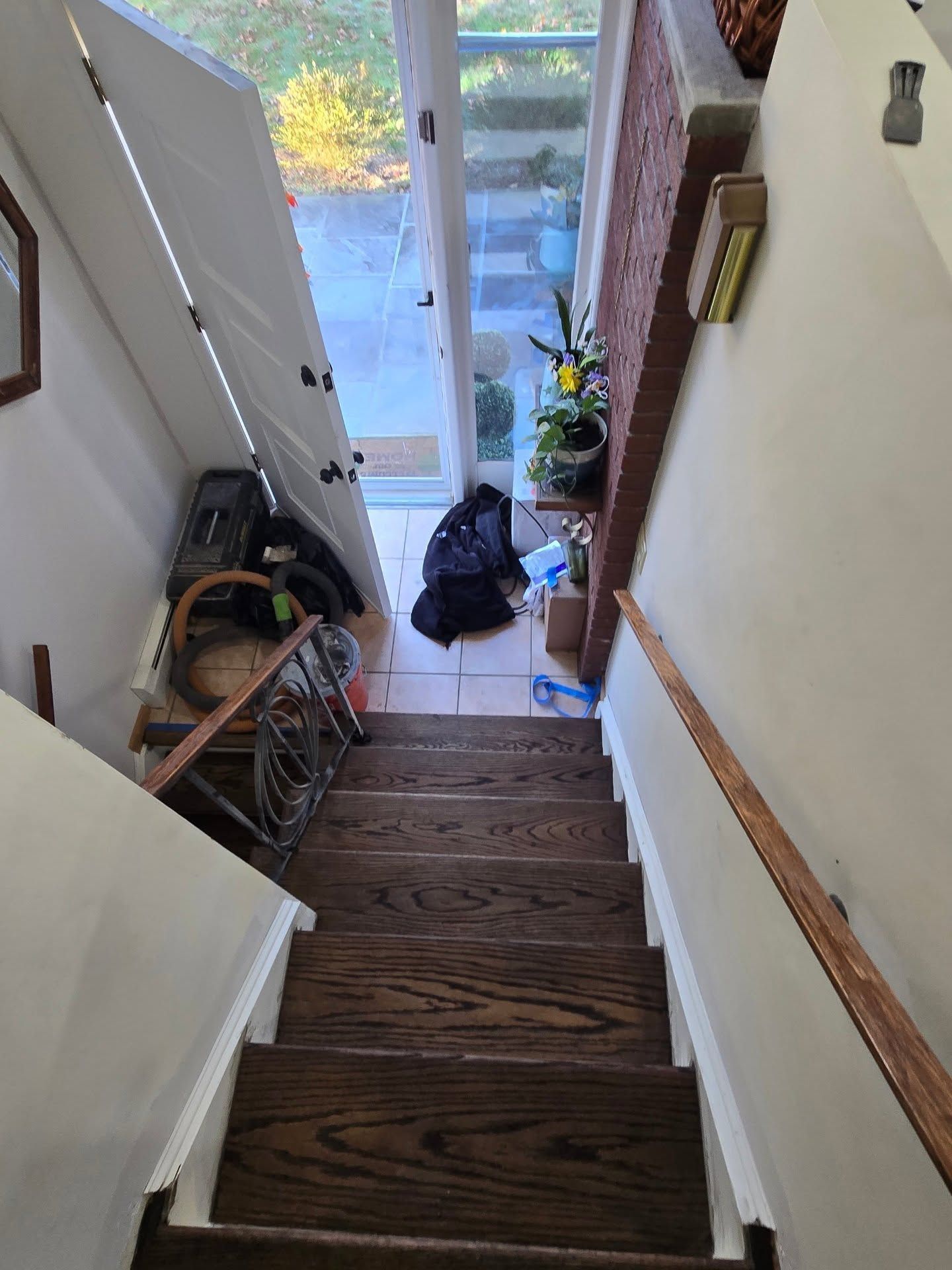 A view looking down a wooden staircase toward an open glass door with scattered items and a small plant on the floor.