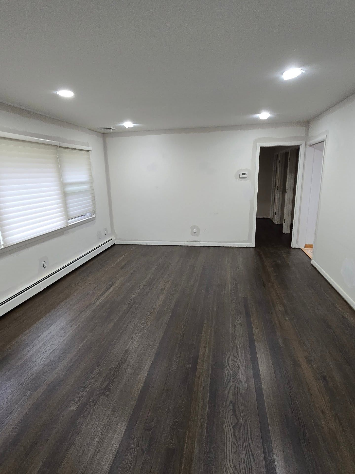 A brightly lit empty room with white walls, recessed ceiling lights, dark wood-look plank flooring, and a large window.