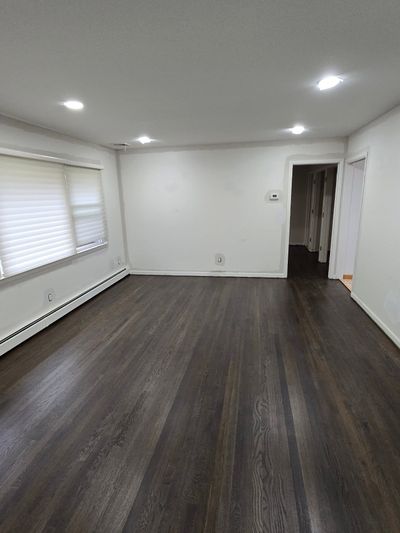 A wide, empty room with white walls, recessed ceiling lights, dark wood-look flooring, and a large window with blinds.