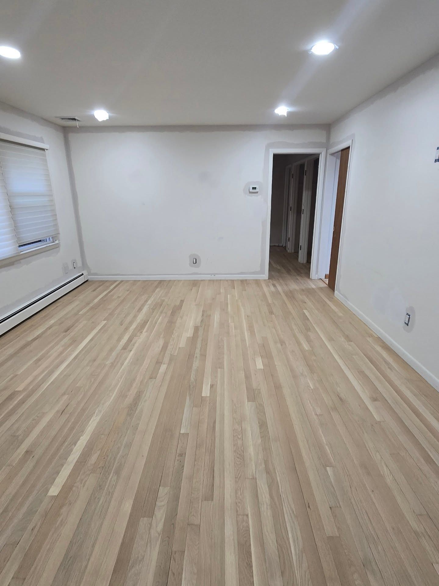 A bright, empty room with light-toned hardwood floors, white walls, recessed ceiling lighting, and a doorway.