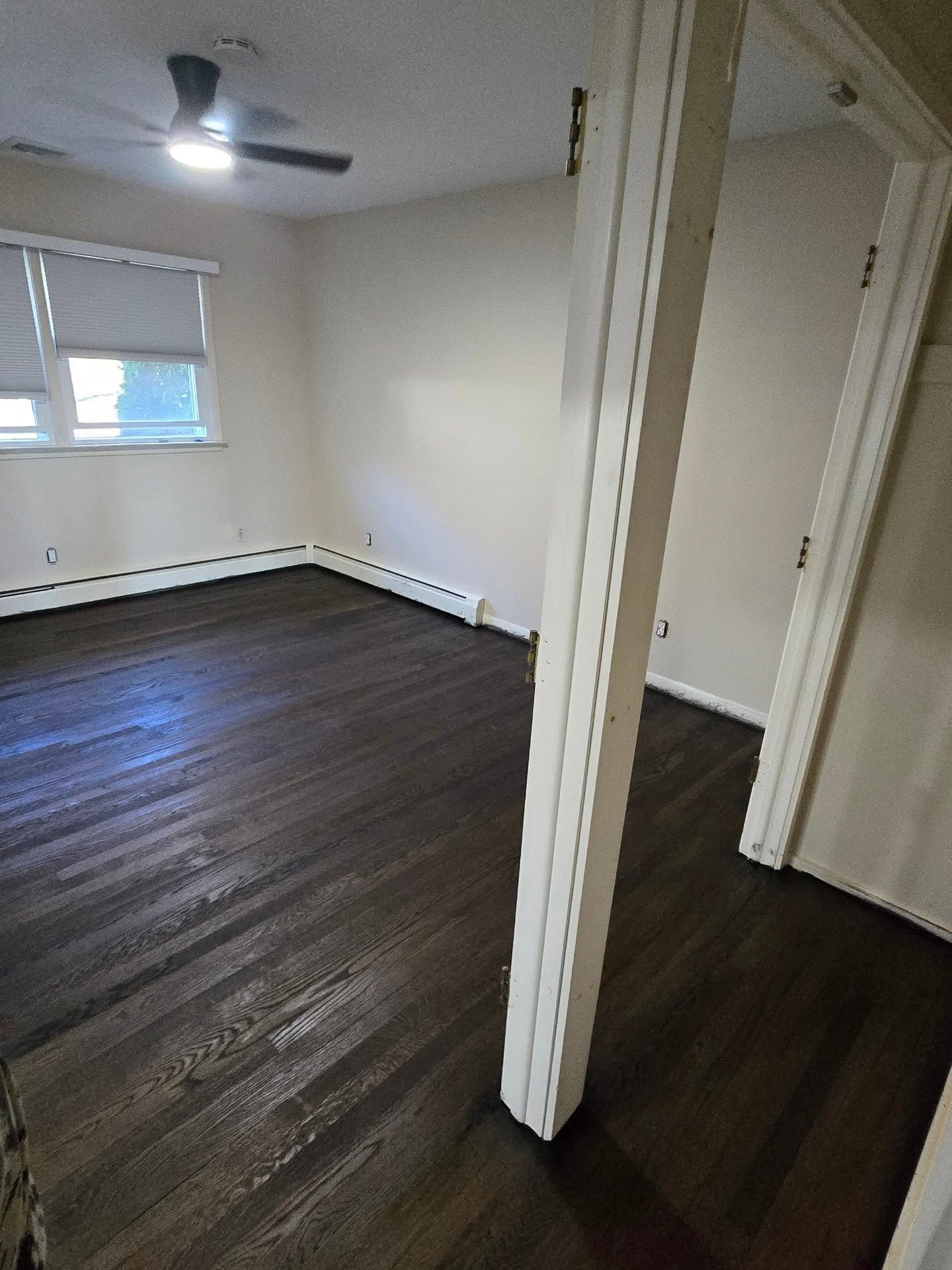 A bright, empty room with dark wood flooring, beige walls, a ceiling fan, and a view of a window through a doorway.