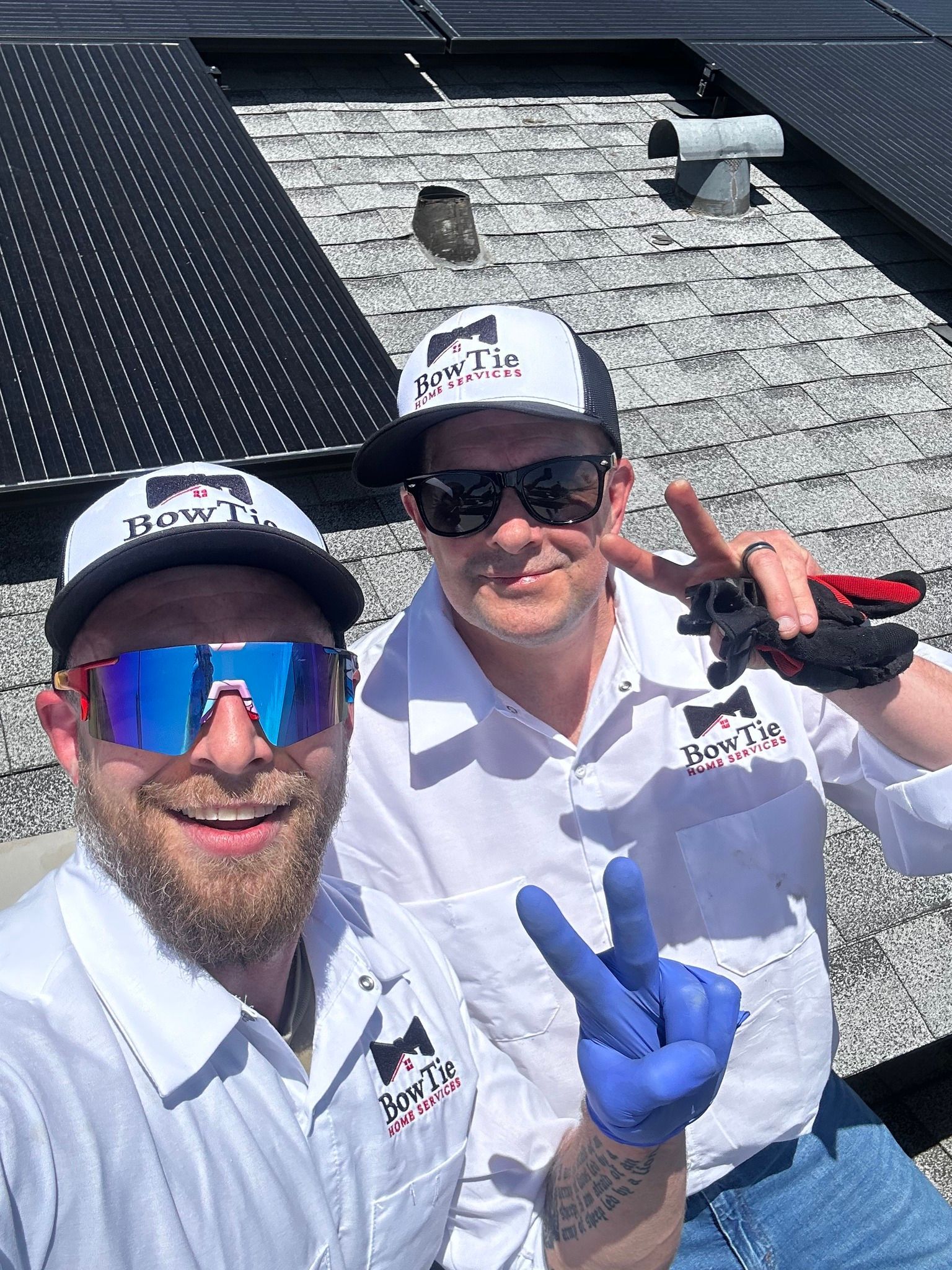 Two people in white uniforms and baseball caps take a selfie on a rooftop with solar panels, both making peace signs.