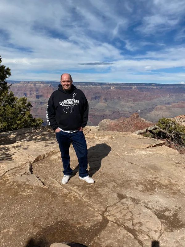 A person stands on a rocky ledge overlooking the Grand Canyon under a partly cloudy blue sky.