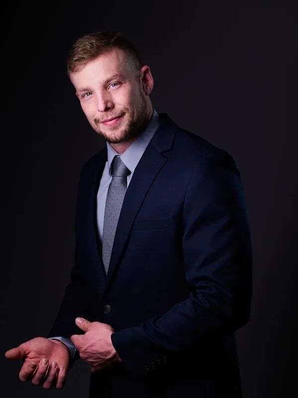 A person wearing a navy blue suit and gray tie stands against a dark background, smiling and gesturing with their hand.