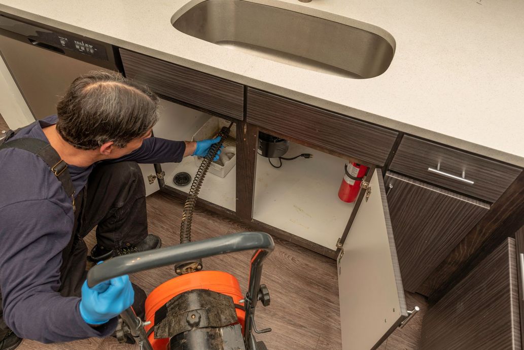 A person in blue gloves operates a plumbing snake to clear a drain under a kitchen sink.