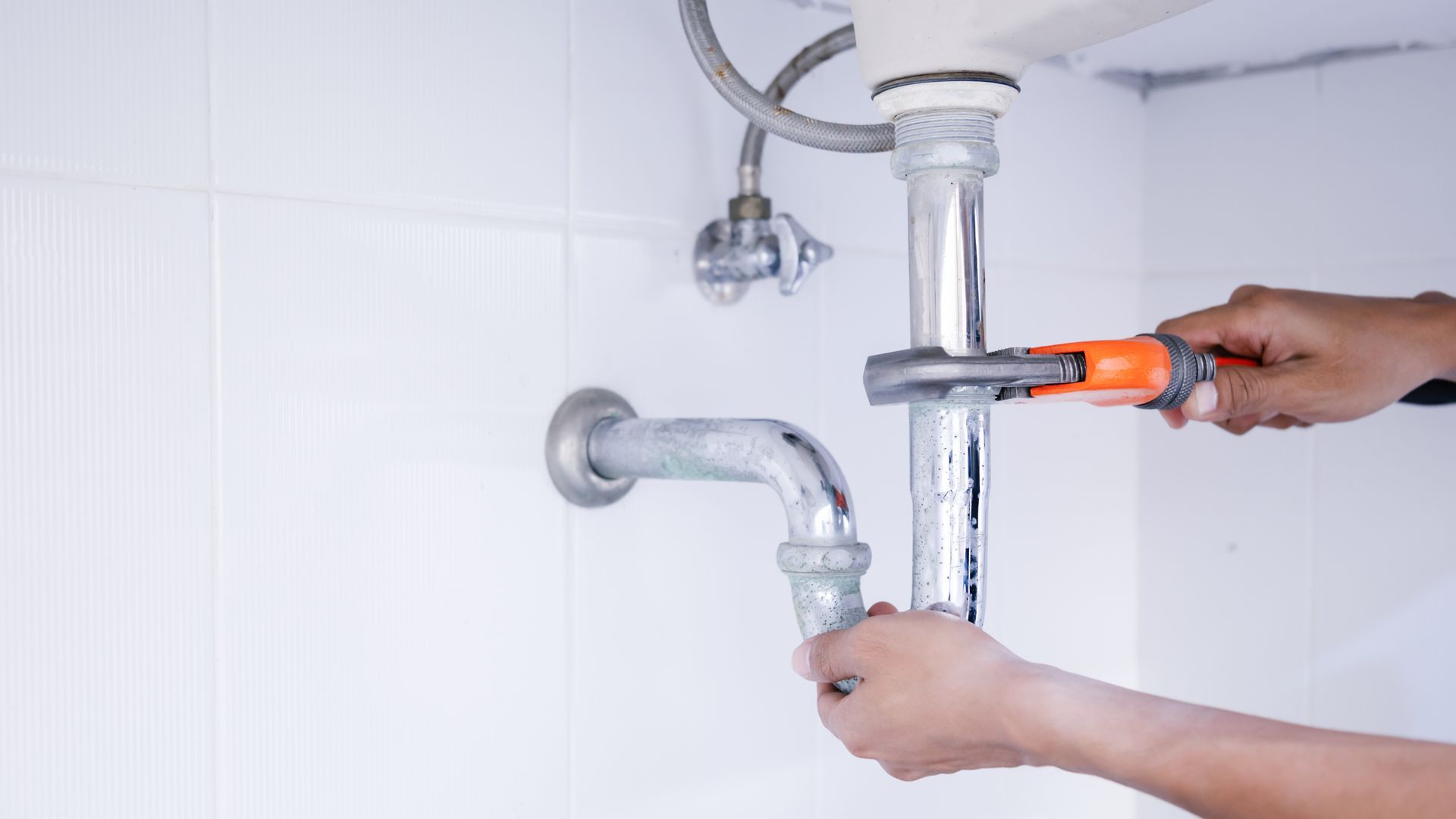 A person uses a wrench to tighten a chrome drain pipe under a sink against a white tiled wall.