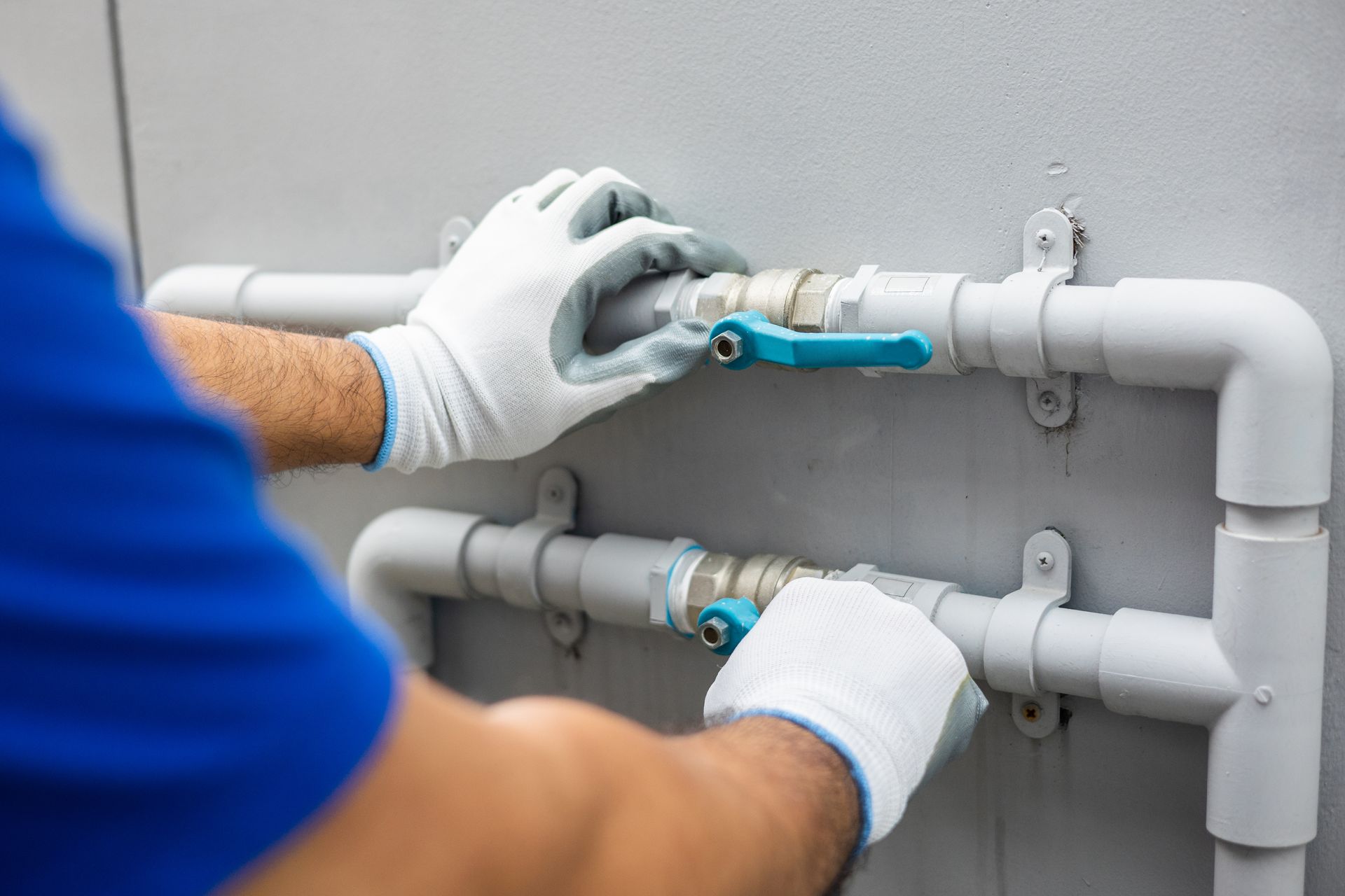A person wearing white work gloves adjusting blue-handled ball valves on gray pipes against a wall.