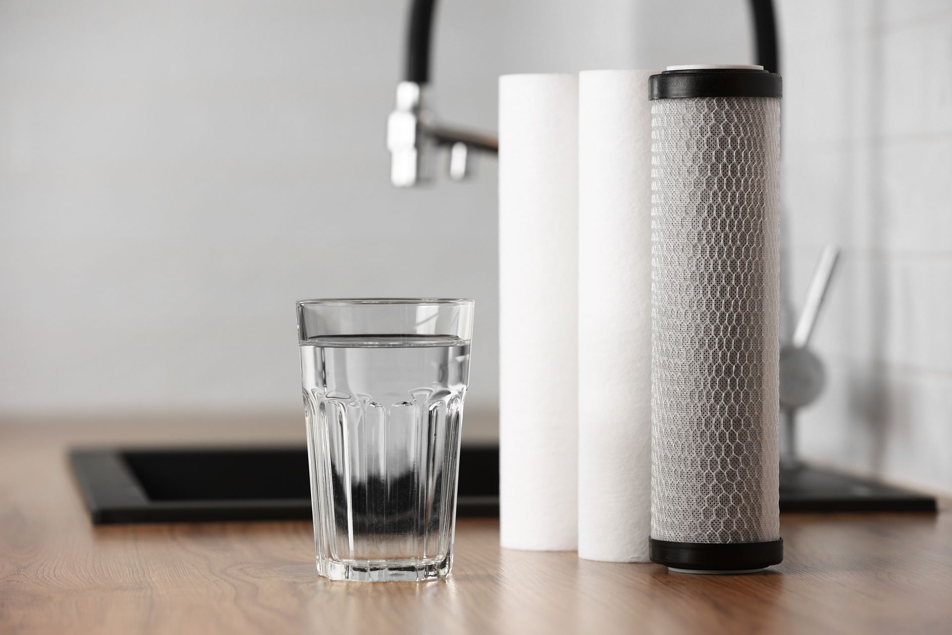 A glass of water stands on a wooden counter next to three cylindrical water filters in front of a kitchen faucet.