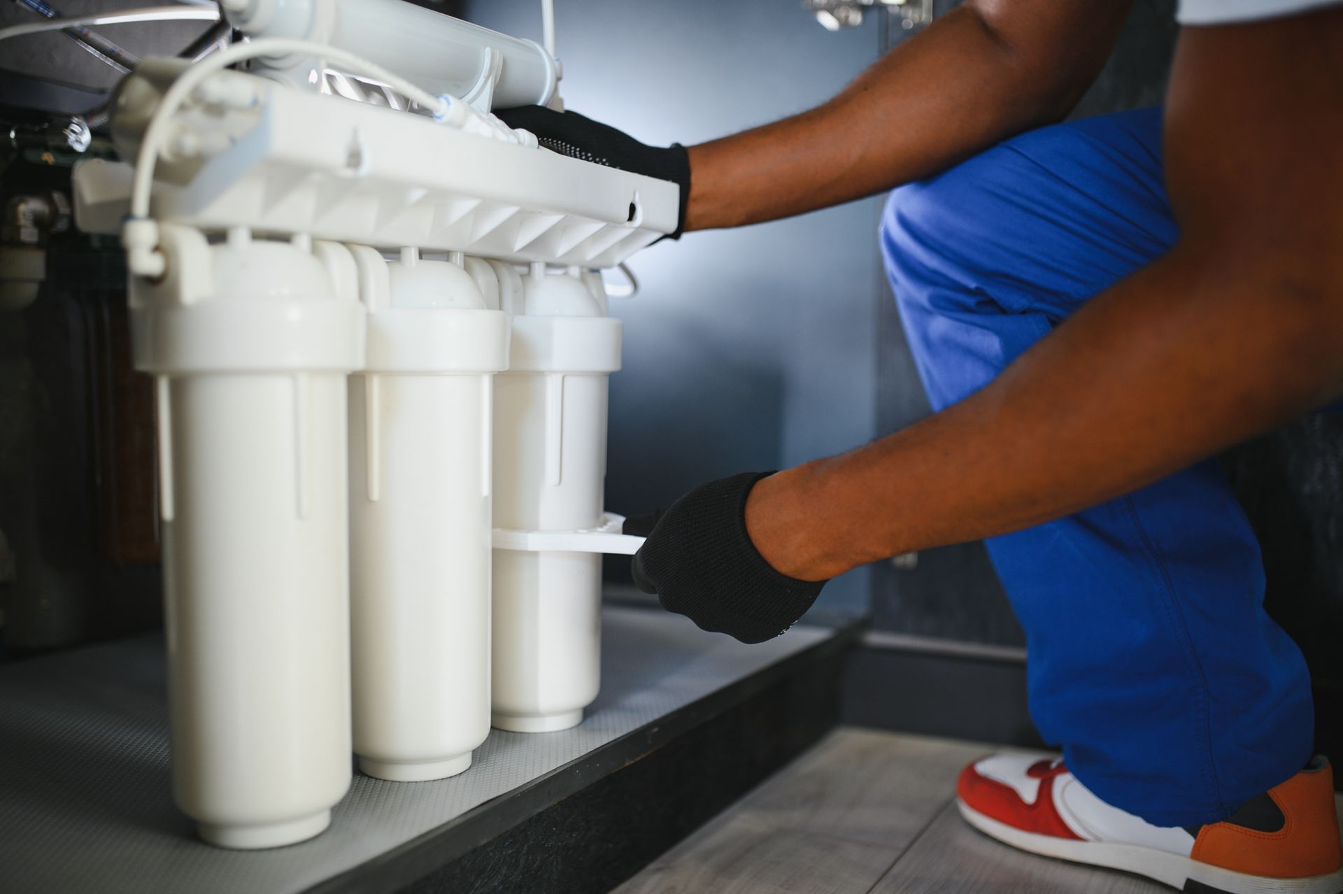 A technician in blue pants and black gloves installs white water filtration cartridges under a kitchen sink.