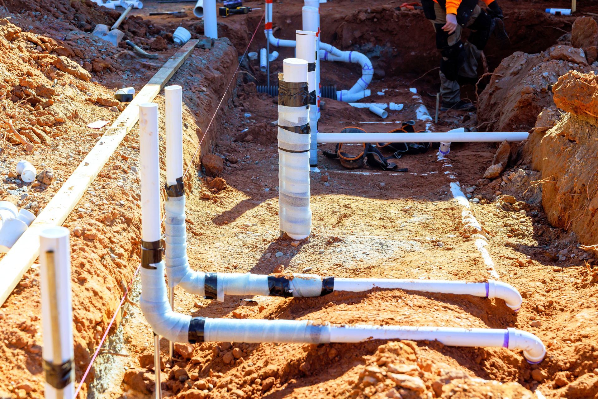 Construction site with white PVC pipes installed in trenches of red dirt, showing ongoing underground plumbing work.