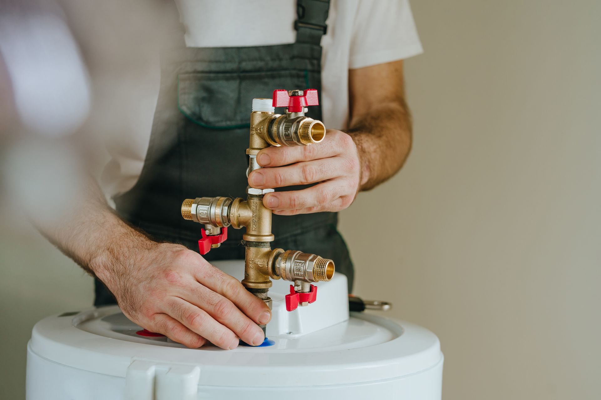 A person in work overalls installs a brass valve assembly onto the top of a white water heater.