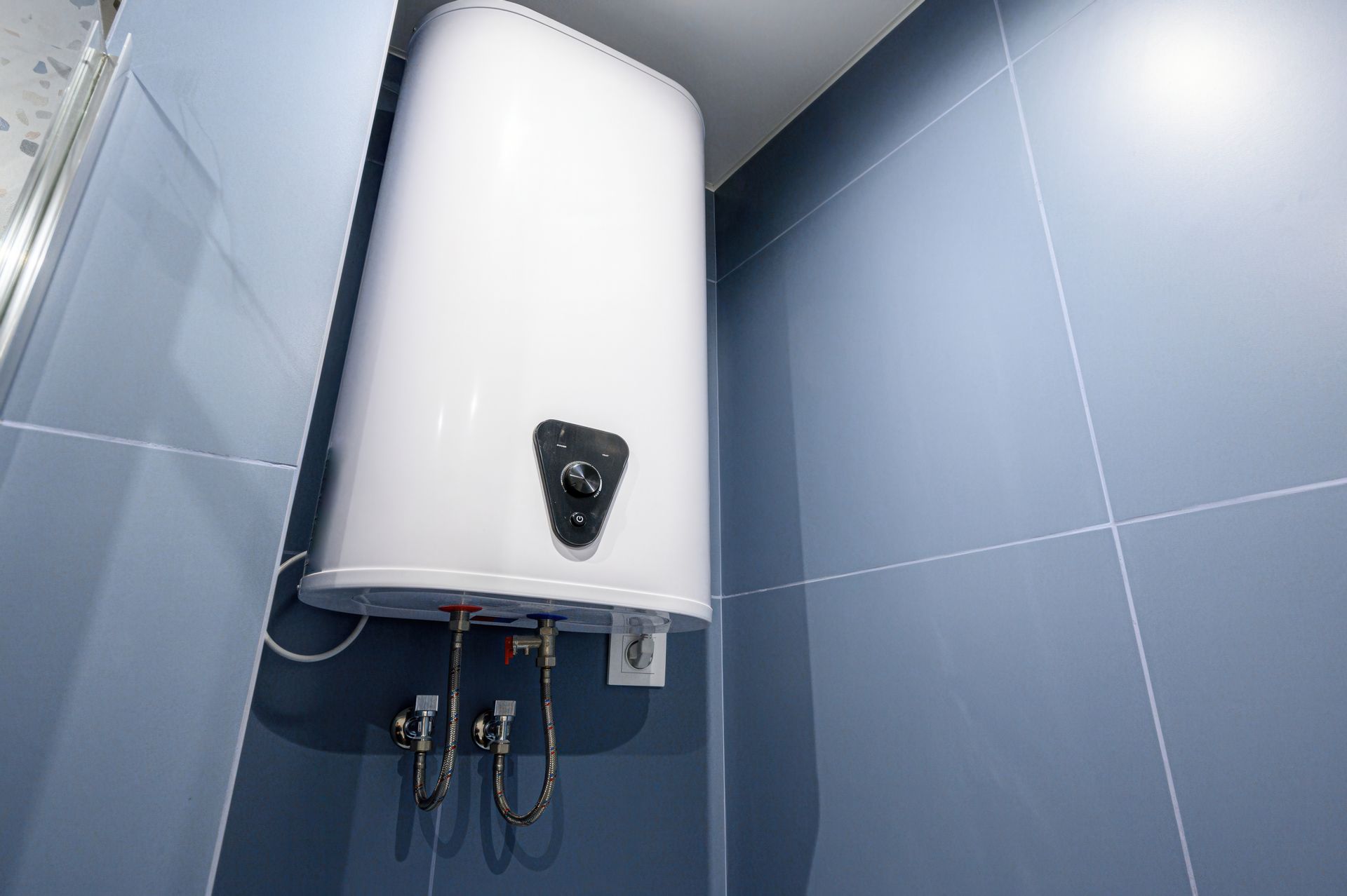 A white electric water heater mounted on a blue tiled wall with exposed pipes and a control dial.