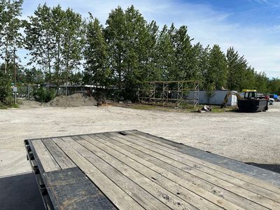 Flatbed trailer in a gravel lot with trees, construction debris, and a small vehicle in the background.