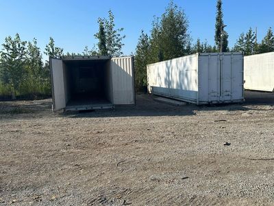 Two white shipping containers on a gravel lot; one has an open door. Green trees in the background.