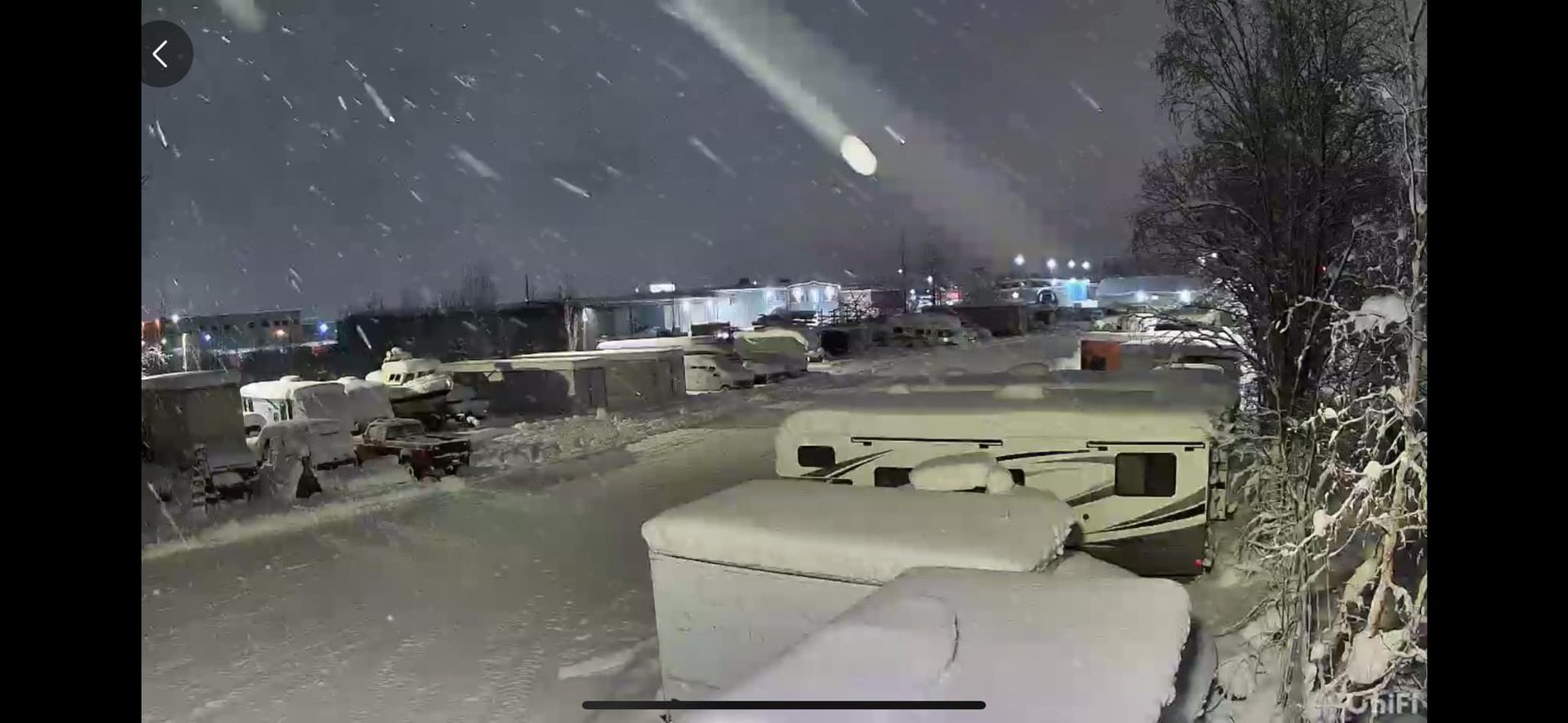 A bright meteor streaking across a snowy sky above a parking lot with covered vehicles and falling snow.