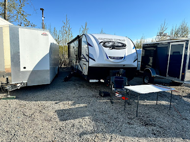 RV and two trailers parked outdoors on gravel with a folding table and camp chair.