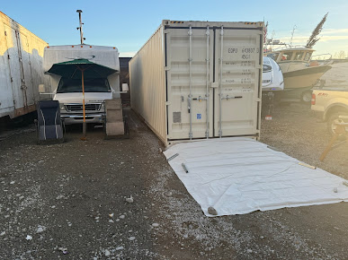 White shipping container with extended white tarp, next to a van under an awning.