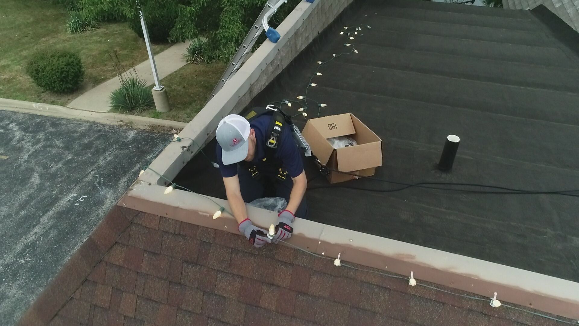 Worker in a hard hat repairs a rooftop edge near a cardboard box and black vent installing christmas lights