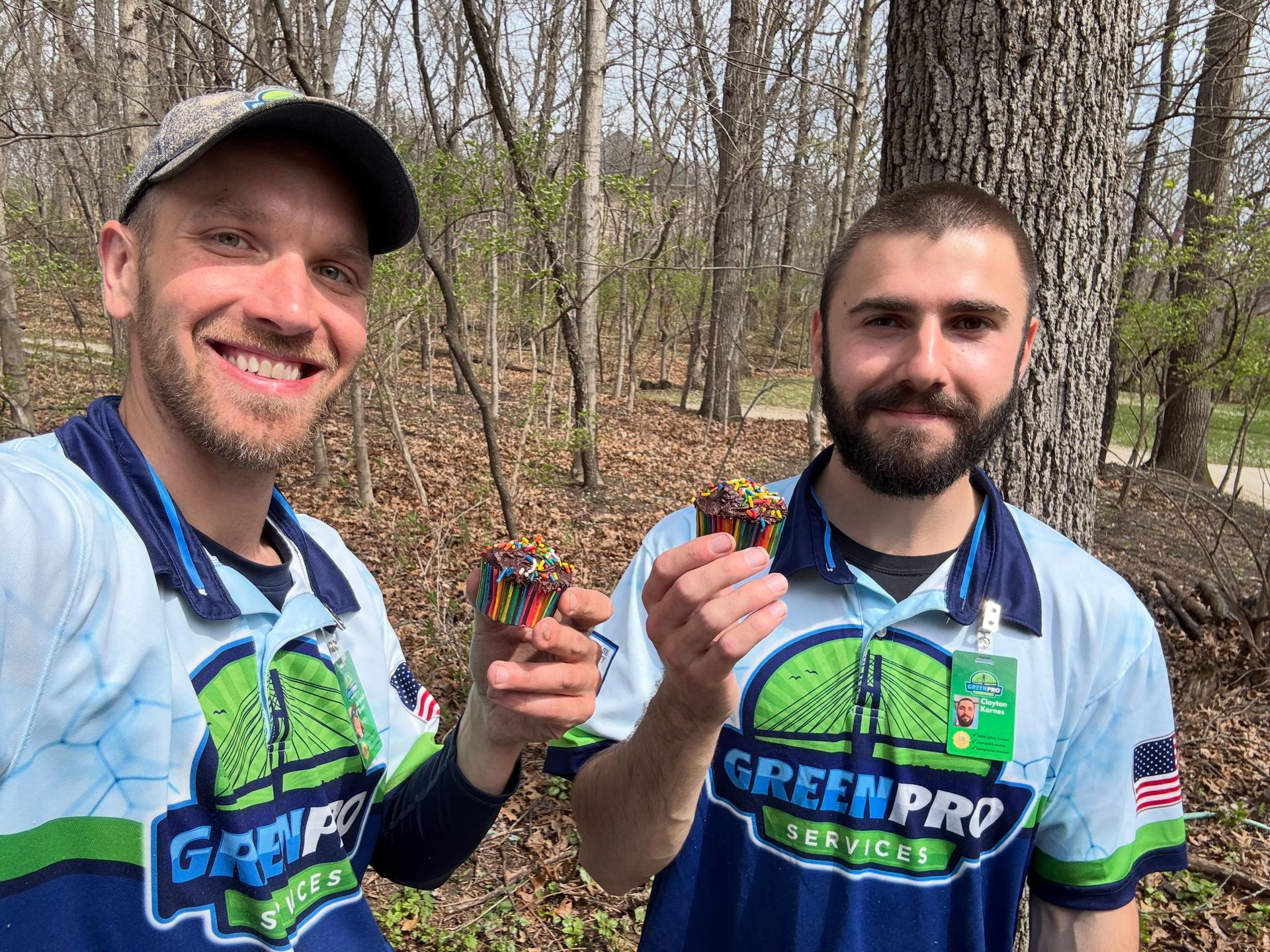 Two men in Green Pro jerseys smiling and holding small colorful items in a wooded area