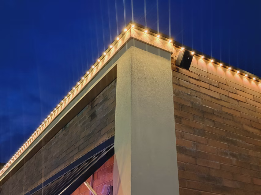 Brick building corner with lit roofline from permanent RGB lighting against a deep blue evening sky