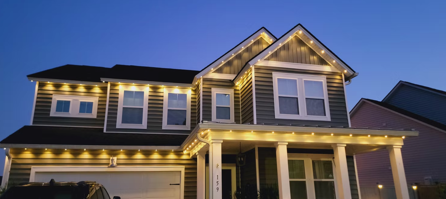 Two-story house at dusk with warm exterior lights and a lit roofline under a blue sky